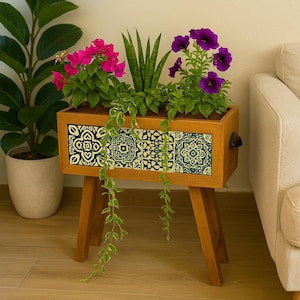 Op de afbeelding: Een houten plantenbak met een decoratief blauw en wit tegelontwerp aan de voorkant. De plantenbak is gevuld met verschillende planten, waaronder roze bougainvillea, paarse petunia's en groen blad. De plantenbak staat op vier houten poten.
