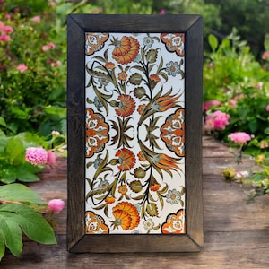 Może przedstawiać: Płytka ceramiczna w ramie, z białym tłem i kwiatowym wzorem w odcieniach pomarańczowego, zielonego i niebieskiego. Płytka jest otoczona ciemną drewnianą ramą.