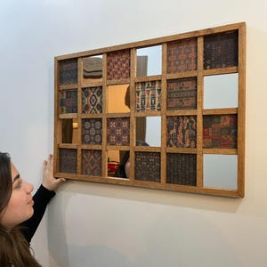 May include: A rectangular wooden framed mirror with a grid of 16 smaller mirrors. Each smaller mirror is framed with a thin wooden border and has a different patterned fabric behind it. The mirror is hanging on a white wall.