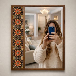 May include: Rectangular wooden framed mirror with a decorative floral panel on the left side. The panel features orange and yellow flowers on a black background. The mirror reflects a room with a chandelier and a person holding a blue phone.