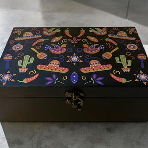 May include: Black wooden box with a hinged lid and a latch closure. The lid is decorated with a colorful pattern of Mexican-inspired designs, including sombreros, cacti, maracas, and chili peppers.