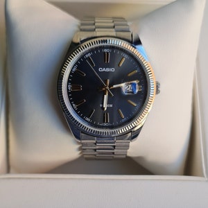 May include: A silver Casio wristwatch with a black face and gold accents. The watch has a date window and a rotating bezel with numbers.