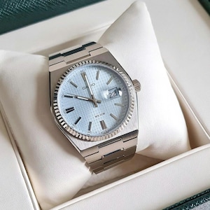May include: A silver Casio wristwatch with a white face and a date window displaying the number 22. The watch has a silver band and a silver bezel with black markings. The watch is in a white box with a green lid.