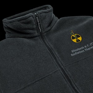 May include: A dark gray fleece jacket with a zipper closure. The jacket has an embroidered logo with a snake wrapped around a yellow radiation symbol and the text "Elisabeth R.T. (T) Radiation Therapist".