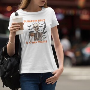 Pode incluir: Camiseta branca com um esqueleto segurando um latte de especiarias de abóbora e o texto "Pumpkin Spice & X-Ray Nights".