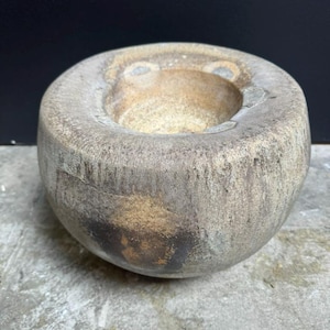 Puede incluir: Un macetero redondo de cerámica gris con una superficie rugosa y texturizada. El macetero tiene una abertura ancha y poco profunda y un fondo ligeramente redondeado.