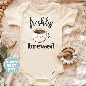 May include: A cream-colored baby onesie with short sleeves features a black graphic of a coffee cup with a smiling face and the text "freshly brewed".