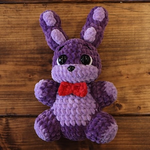 以下が含まれることがあります： 大きな黒い目と赤い蝶ネクタイが付いた、手作りの紫色のウサギのぬいぐるみ。ウサギは、より明るい紫色の斑点のある長い耳を持ち、柔らかく質感のある素材で作られています。おもちゃは木製の表面に置かれています。