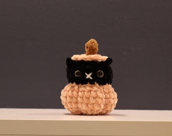 May include: A handmade crochet plushie in the shape of a small creature. The plushie has a peach-colored body, a black head with white cross-shaped mouth, and a small brown top. The eyes are black.
