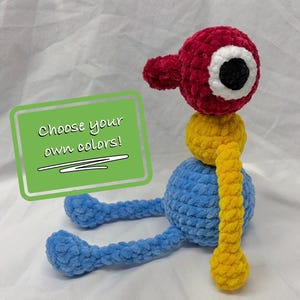 May include: A plush crochet toy featuring a red head with a large white and black eye, a yellow midsection, and a blue spherical base with long blue legs and a yellow arm. A green rectangular sign displays the text Choose your own colors!