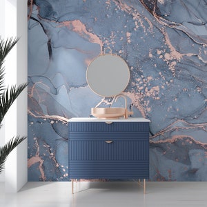 May include: A blue vanity cabinet with two drawers and gold metal legs. The cabinet has a white countertop with a copper sink and a round gold mirror above it. The background is a blue and gold marble wallpaper.