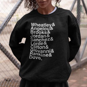 May include: Black sweatshirt with white text listing names: Wheatley, Angelou, Brooks, Jordan, Sanchez, Lorde, Clifton, Giovanni, Rankine, and Dove. The sweatshirt is a crew neck style.