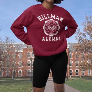 Pode incluir: Uma camisa de moletom vinho com um brasão branco com o texto "Hillman Alumni" e um selo com o texto "Deus Non Domdum Te Confecit 1881".