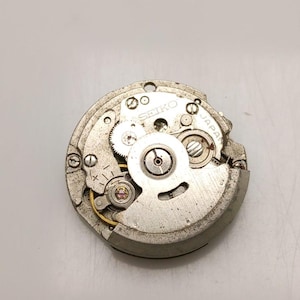 K&ouml;nnte beinhalten: Nahaufnahme eines Seiko-Uhrwerks. Der silberfarbene Mechanismus verf&uuml;gt &uuml;ber Zahnr&auml;der, Schrauben und das Wort "SEIKO" auf der Oberfl&auml;che. Die Worte "JAPAN" sind ebenfalls sichtbar.
