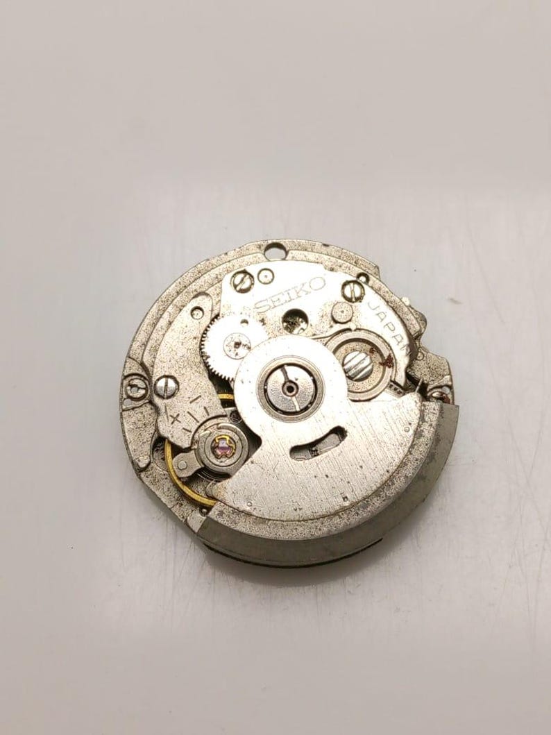 K&ouml;nnte beinhalten: Nahaufnahme eines Seiko-Uhrwerks. Der silberfarbene Mechanismus verf&uuml;gt &uuml;ber Zahnr&auml;der, Schrauben und das Wort "SEIKO" auf der Oberfl&auml;che. Die Worte "JAPAN" sind ebenfalls sichtbar.