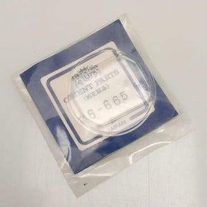 May include: A clear, circular watch crystal in a plastic bag, resting on a blue and white paper. The paper has the text "ORIENT PARTS" and "6-665" printed on it. The packaging is transparent, allowing the crystal to be visible.