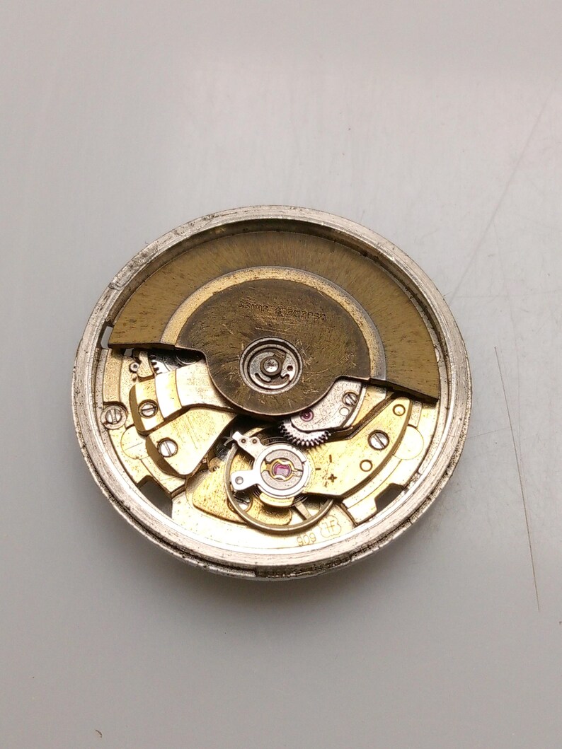 May include: A detailed view of a watch movement, showcasing its complex internal mechanisms. The components are predominantly gold and silver, arranged in a circular pattern. The image highlights the precision engineering of the watch.