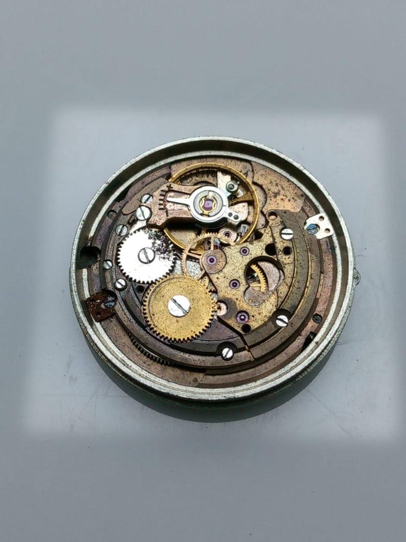 May include: Close-up of a disassembled watch mechanism. The intricate internal components include various gears, springs, and other small parts. The watch's metal casing is visible, surrounding the internal workings. The gears are gold and silver in color.