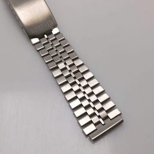 Könnte beinhalten: Ein silberfarbenes Citizen-Uhrenarmband. Das Armband besteht aus kleinen, quadratischen Metallgliedern. Der rechteckige Verschluss trägt die Gravur "CITIZEN". Das Armband ist verstellbar und passt für verschiedene Handgelenkgrößen.