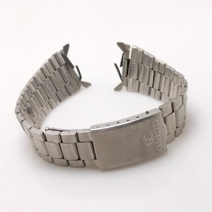 Pode incluir: Uma pulseira de relógio de metal prateado com fecho dobrável. A pulseira é feita de elos retangulares e apresenta o logotipo Seiko no fecho. A pulseira está solta em ambas as extremidades.