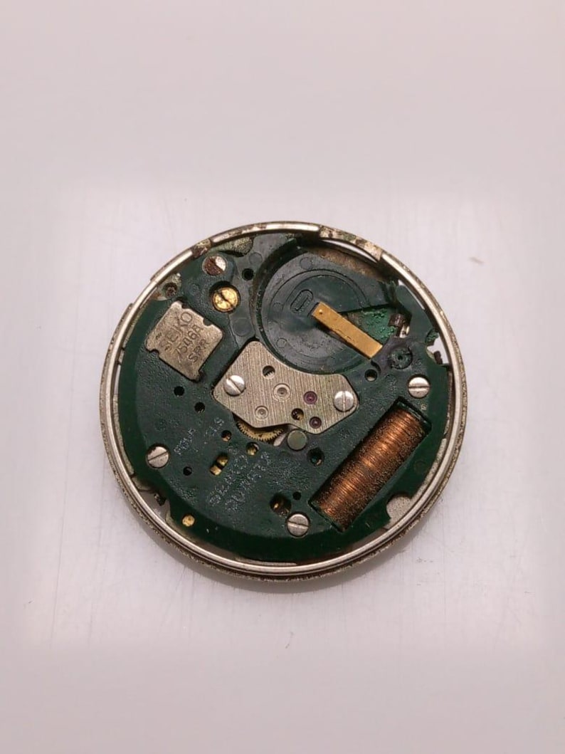 May include: A disassembled watch movement with a green circuit board and metallic components. The watch movement has a silver metal casing. The text "SEIKO 7506A SPR" is visible on the circuit board.
