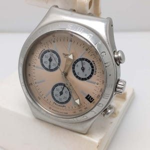 May include: A silver-toned Swatch chronograph watch with a peach-colored face. The watch features multiple subdials, a date display, and a bezel with markings. The watch is mounted on a white stand.