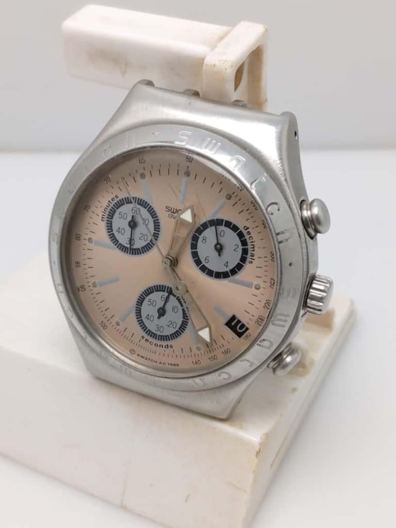 May include: A silver-toned Swatch chronograph watch with a peach-colored face. The watch features multiple subdials, a date display, and a bezel with markings. The watch is mounted on a white stand.