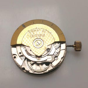 May include: A close-up of a watch movement with visible gears and components. The gold-colored rotor is marked "RADO 25 JEWELS". A small, textured crown is attached. The watch mechanism is silver and gold in color.