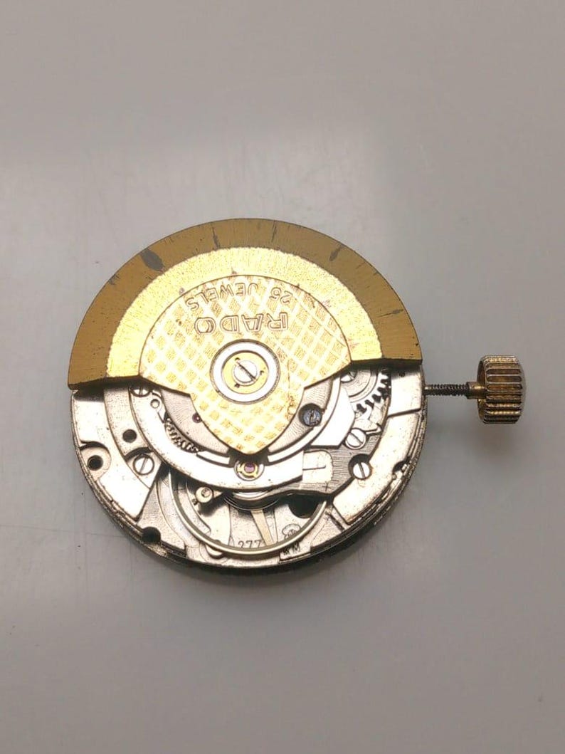 May include: A close-up of a watch movement with visible gears and components. The gold-colored rotor is marked "RADO 25 JEWELS". A small, textured crown is attached. The watch mechanism is silver and gold in color.