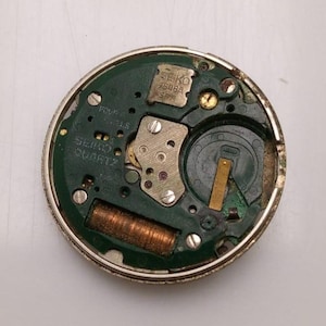May include: A close-up of a Seiko watch movement. The watch movement has a green circuit board with various components, including a battery, gears, and a coil. The text "SEIKO 7506A SPR" is visible. The watch movement is circular and has a silver metal casing.