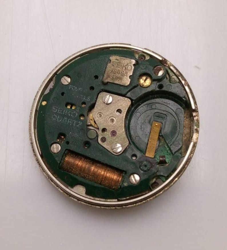 May include: A close-up of a Seiko watch movement. The watch movement has a green circuit board with various components, including a battery, gears, and a coil. The text "SEIKO 7506A SPR" is visible. The watch movement is circular and has a silver metal casing.