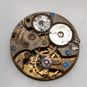 May include: Close-up of a vintage watch movement with intricate gears and components. The metal parts are in shades of gold, silver, and bronze, with blue accents. The text "LONGINES 6786472" is visible on the movement.