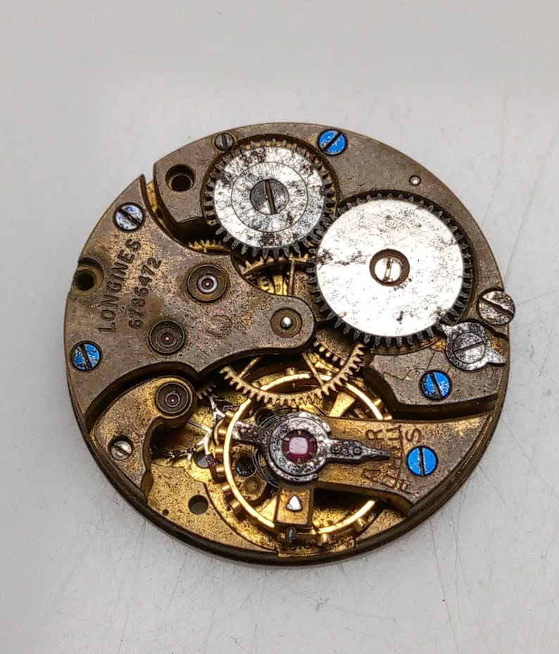 May include: Close-up of a vintage watch movement with intricate gears and components. The metal parts are in shades of gold, silver, and bronze, with blue accents. The text "LONGINES 6786472" is visible on the movement.