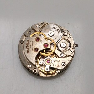 May include: Close-up of a watch movement with intricate gears and components. The watch mechanism features gold-colored gears, red jewels, and the text "ROAMER 17 JEWELS SWISS MADE". The watch movement is circular and made of metal.