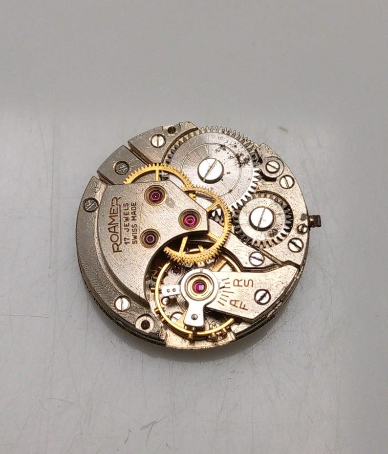 May include: Close-up of a watch movement with intricate gears and components. The watch mechanism features gold-colored gears, red jewels, and the text "ROAMER 17 JEWELS SWISS MADE". The watch movement is circular and made of metal.