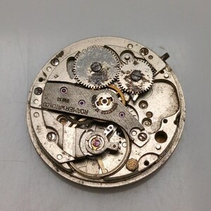 May include: Close-up of a vintage watch movement with gears, screws, and the text "ROAMER WATCH CO SWISS". The watch mechanism is circular and made of metal, with visible jewels and intricate details.