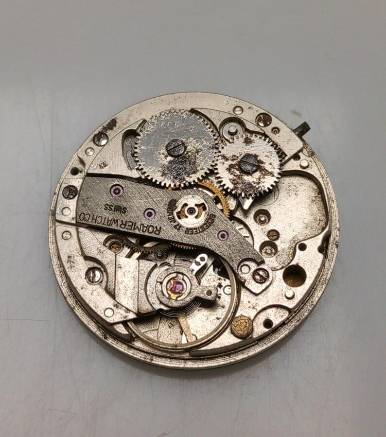 May include: Close-up of a vintage watch movement with gears, screws, and the text "ROAMER WATCH CO SWISS". The watch mechanism is circular and made of metal, with visible jewels and intricate details.