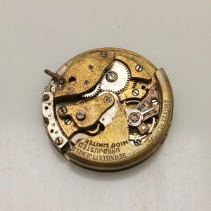 Puede incluir: Un movimiento de reloj vintage con engranajes, tornillos y joyas visibles. El mecanismo dorado está marcado con "SEVENTEEN 17 JEWELS UNADJUSTED MIDO LIMITED SWITZERLAND". El movimiento del reloj es circular.