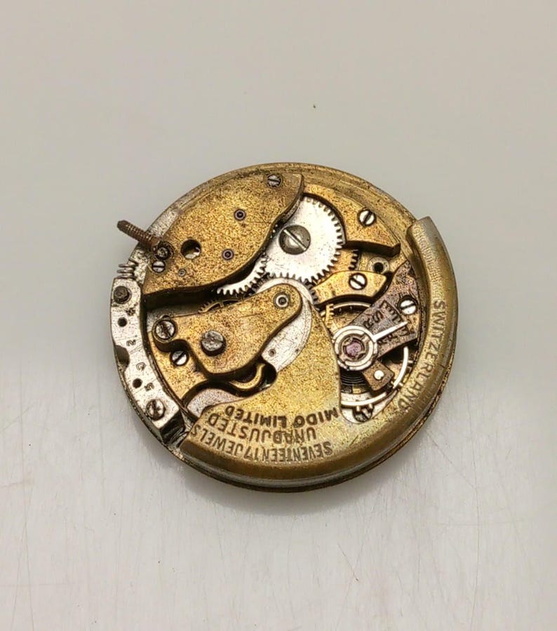 Może przedstawiać: Zabytkowy mechanizm zegarka z widocznymi kołami zębatymi, śrubami i kamieniami. Złoty mechanizm jest oznaczony napisem "SEVENTEEN 17 JEWELS UNADJUSTED MIDO LIMITED SWITZERLAND". Mechanizm zegarka jest okrągły.