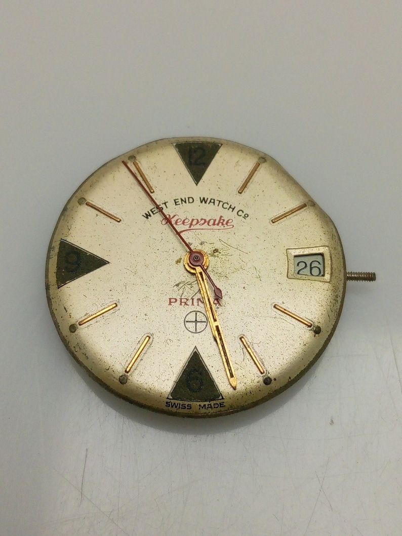 Puede incluir: Una esfera de reloj vintage con agujas y marcadores dorados. La esfera es blanca con marcas negras y el texto "West End Watch Co. Keepsake Prima Swiss Made" y el n&uacute;mero "26".