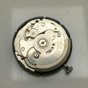 May include: Close-up of a Seiko watch movement, showcasing intricate gears, springs, and jewels. The silver-toned components are set against a dark background. The text "SEIKO TIME TWENTY-THREE JEWELS JAPAN" is visible.
