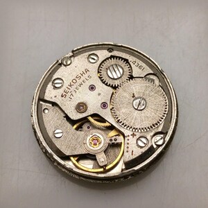 May include: A detailed view of a vintage watch mechanism, featuring silver and gold metal components, gears, and screws. The text "SEIKOSHA 17 JEWELS" is visible. The circular mechanism has small red jewels.