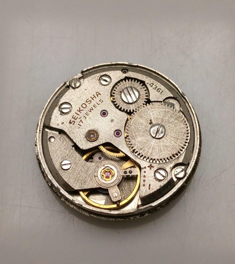 May include: A detailed view of a vintage watch mechanism, featuring silver and gold metal components, gears, and screws. The text "SEIKOSHA 17 JEWELS" is visible. The circular mechanism has small red jewels.