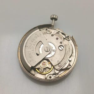 Pode incluir: Uma aproxima&ccedil;&atilde;o de um movimento de rel&oacute;gio Orient 21J 46943 Japan prateado. O mecanismo do rel&oacute;gio &eacute; redondo com v&aacute;rias engrenagens, parafusos e uma haste de corda. O texto "ORIENT 21J 46943 JAPAN" &eacute; vis&iacute;vel.