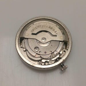 May include: A silver-toned watch movement with intricate gears and components. The text "ORIENT 21 JEWELS 46941" is visible on the face. A small, textured crown is on the side. The watch mechanism is circular.