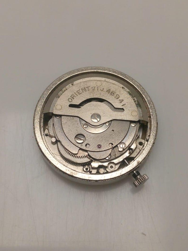 May include: A silver-toned watch movement with intricate gears and components. The text "ORIENT 21 JEWELS 46941" is visible on the face. A small, textured crown is on the side. The watch mechanism is circular.