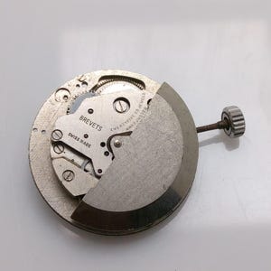 May include: A silver-toned watch movement, partially disassembled, showing intricate gears and screws. The text "BREVETS SWISS MADE" is visible. A winding stem with a crown is attached to the mechanism.