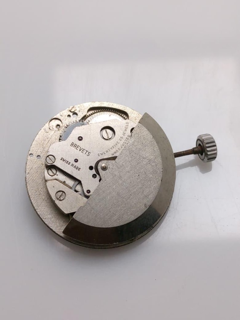 May include: A silver-toned watch movement, partially disassembled, showing intricate gears and screws. The text "BREVETS SWISS MADE" is visible. A winding stem with a crown is attached to the mechanism.