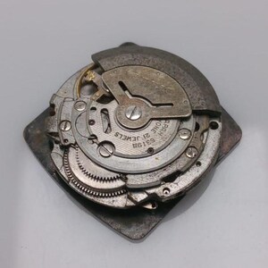 Puede incluir: Mecanismo de reloj desmontado, con engranajes, muelles y otros componentes internos visibles. Las piezas met&aacute;licas son plateadas y grises, con el texto "21 JEWELS" y "JAPAN ONE". Montado sobre una base cuadrada.