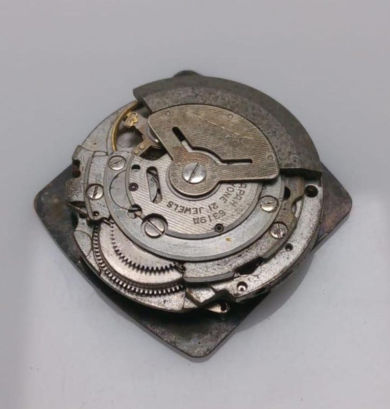 Puede incluir: Mecanismo de reloj desmontado, con engranajes, muelles y otros componentes internos visibles. Las piezas met&aacute;licas son plateadas y grises, con el texto "21 JEWELS" y "JAPAN ONE". Montado sobre una base cuadrada.